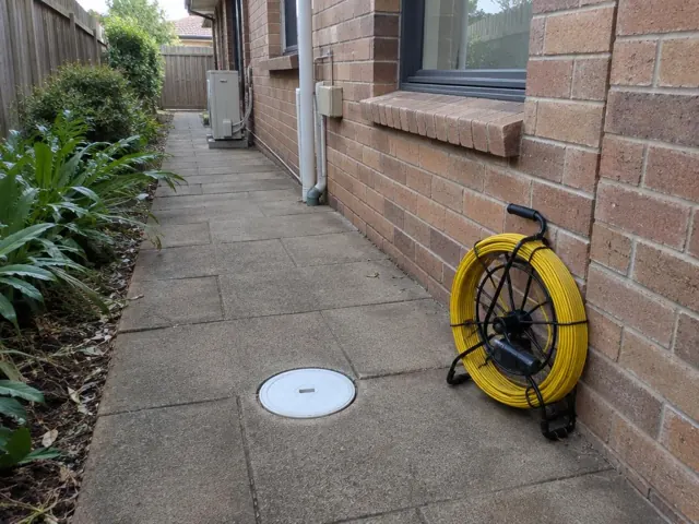 Capped inspection opening on a paved side path beside a brick house, with a coiled camera reel in Macquarie Park