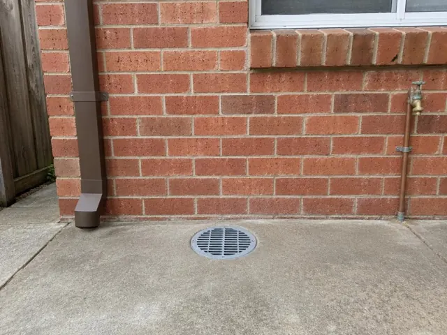 Outdoor concrete gully grate beside a brick wall and downpipe, area clean and dry in West Ryde