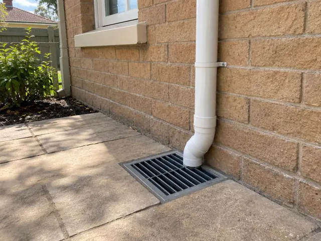 Cleared stormwater grate beside a brick house wall and downpipe on a dry paved path in Ryde